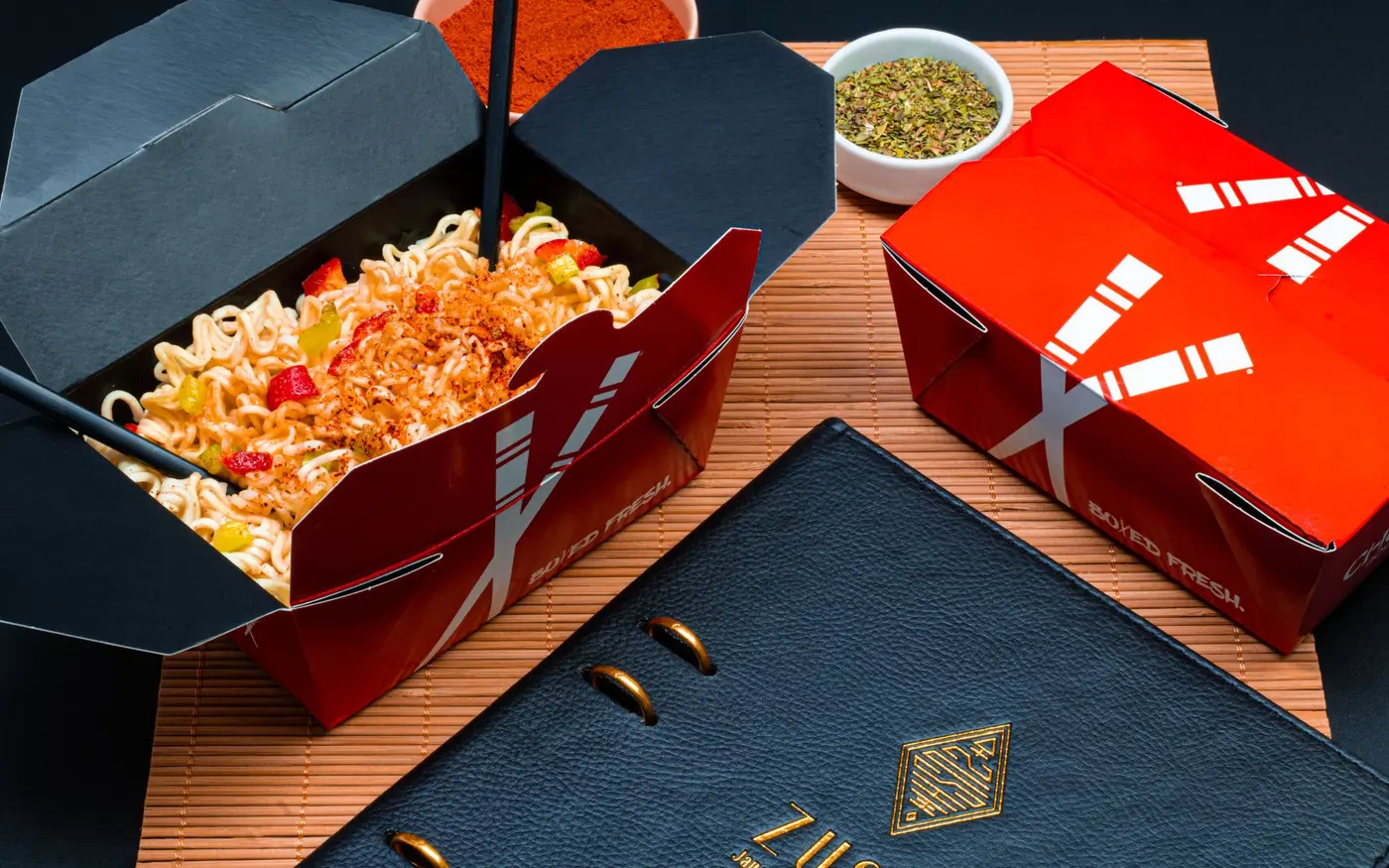 Asian noodles packed into takeout boxes with chopsticks and spices beside a menu