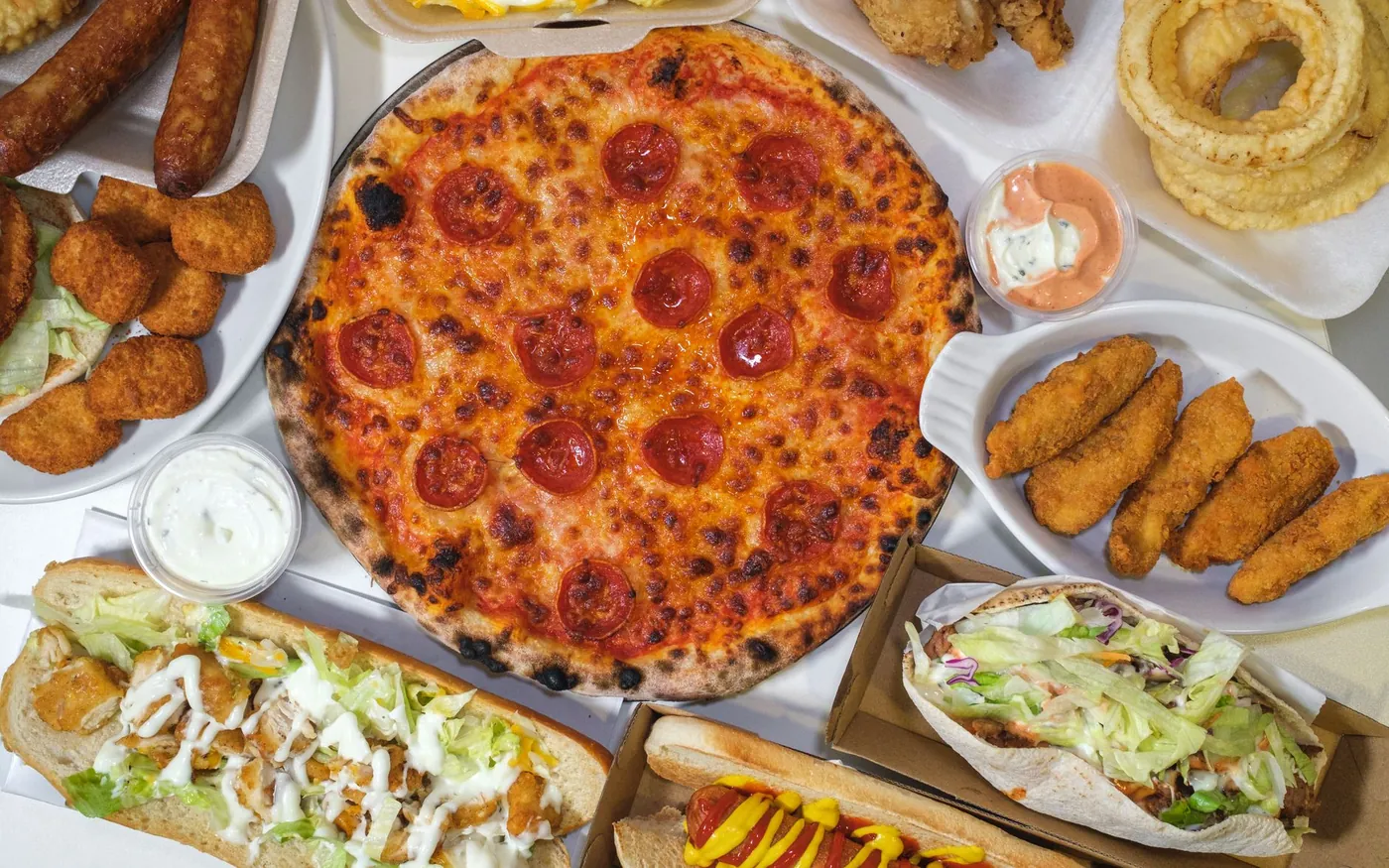 Pizza, subs, hot dogs, wraps, fried chicken tenders and onion rings spread out in takeaway boxes and trays